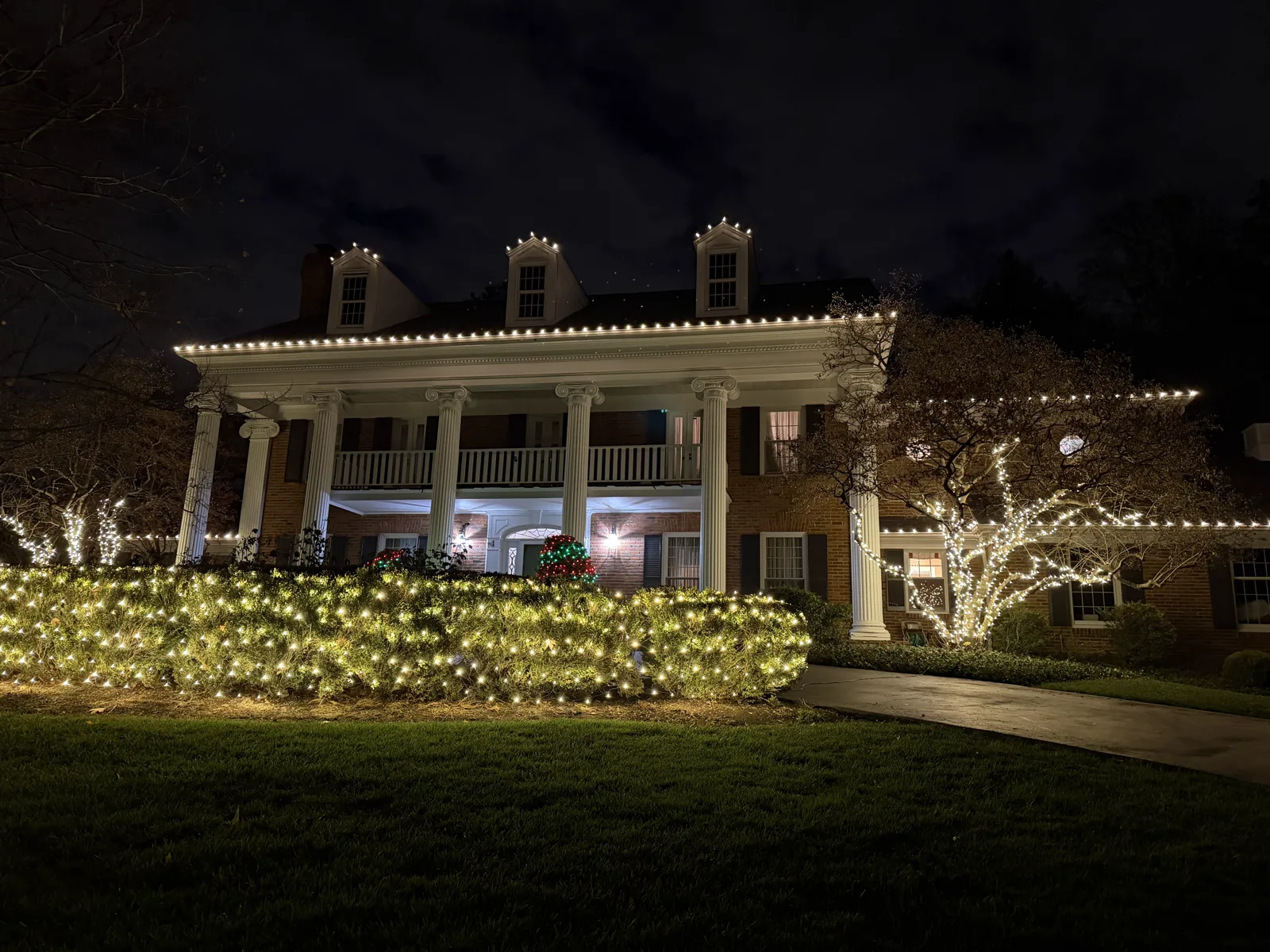 Warm-white roofline lighting with net-lit hedges and wrapped trees on a grand brick colonial