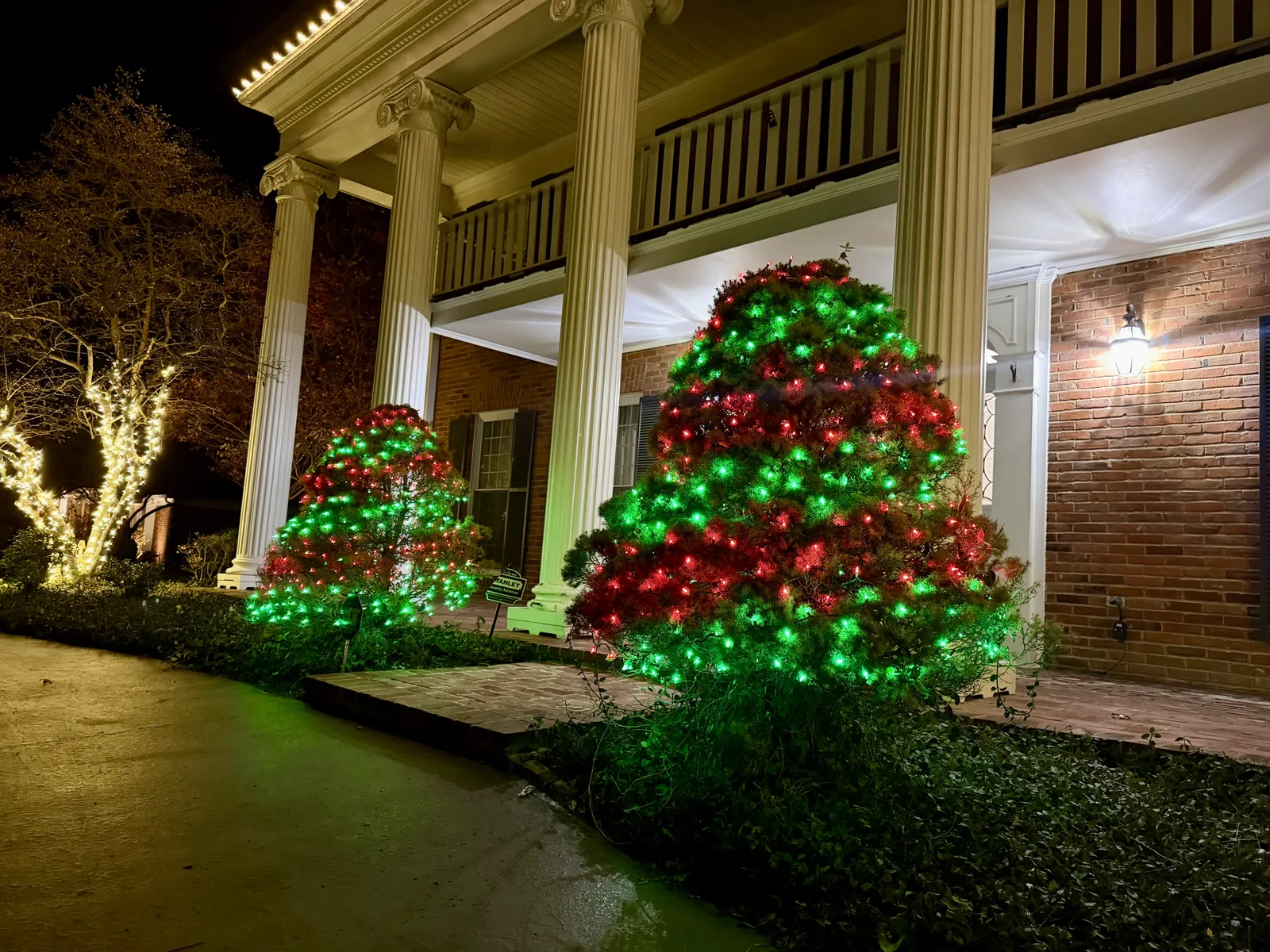 Red and green net lighting on shrubs beside a warm-white wrapped tree at a columned home